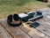 A pair of black headphones, a music production controller, a black portable electronic device with a yellow cable, and a handheld video recorder are placed on a wooden table outdoors. The scene features grassy surroundings.