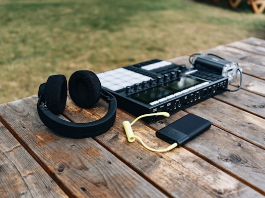 A pair of black headphones, a music production controller, a black portable electronic device with a yellow cable, and a handheld video recorder are placed on a wooden table outdoors. The scene features grassy surroundings.
