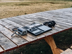 Audio equipment rests on a wooden picnic table outdoors. This includes a set of headphones, an audio mixer or controller with multiple dials and buttons, and a recording device or portable speaker with attached cables. The table is positioned on grass and surrounded by a dry, brown landscape.