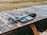 Audio equipment rests on a wooden picnic table outdoors. This includes a set of headphones, an audio mixer or controller with multiple dials and buttons, and a recording device or portable speaker with attached cables. The table is positioned on grass and surrounded by a dry, brown landscape.