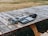 Audio equipment rests on a wooden picnic table outdoors. This includes a set of headphones, an audio mixer or controller with multiple dials and buttons, and a recording device or portable speaker with attached cables. The table is positioned on grass and surrounded by a dry, brown landscape.
