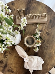 A wooden table with a rustic aesthetic hosts a small arrangement of white flowers in a white vase, a cup of coffee, a neatly folded napkin with a white floral decoration, and a decorative sign that spells 'Home' in metallic letters.