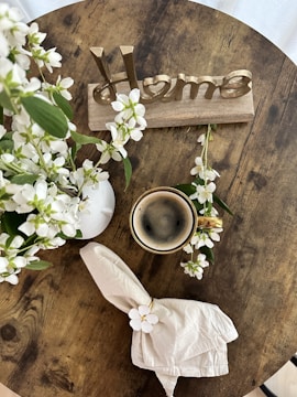 A wooden table with a rustic aesthetic hosts a small arrangement of white flowers in a white vase, a cup of coffee, a neatly folded napkin with a white floral decoration, and a decorative sign that spells 'Home' in metallic letters.