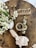 A wooden table with a rustic aesthetic hosts a small arrangement of white flowers in a white vase, a cup of coffee, a neatly folded napkin with a white floral decoration, and a decorative sign that spells 'Home' in metallic letters.