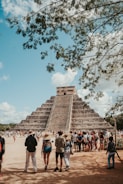 A large stone pyramid with multiple tiers stands prominently in the background, surrounded by a large group of people. Many of the visitors are wearing hats and casual summer clothing, indicating a tourist setting. The pyramid is partially obscured at the top by a tree with overhanging branches. The sky is clear with only a few scattered clouds. The scene conveys a sense of awe and historical significance, with tourists gathered to observe the ancient structure.