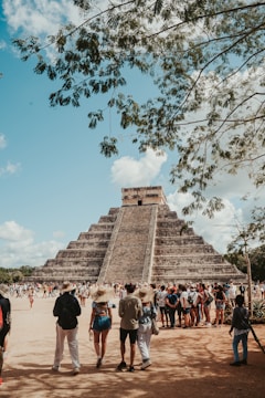 A large stone pyramid with multiple tiers stands prominently in the background, surrounded by a large group of people. Many of the visitors are wearing hats and casual summer clothing, indicating a tourist setting. The pyramid is partially obscured at the top by a tree with overhanging branches. The sky is clear with only a few scattered clouds. The scene conveys a sense of awe and historical significance, with tourists gathered to observe the ancient structure.