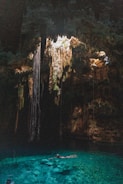 A natural underground pool, known as a cenote, is surrounded by rock formations and illuminated by sunlight streaming through an opening above. Long roots or stalactites hang from the ceiling, creating an otherworldly atmosphere. The water is clear and vibrant turquoise, with a person swimming leisurely in its depths.