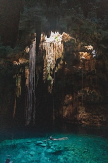 A natural underground pool, known as a cenote, is surrounded by rock formations and illuminated by sunlight streaming through an opening above. Long roots or stalactites hang from the ceiling, creating an otherworldly atmosphere. The water is clear and vibrant turquoise, with a person swimming leisurely in its depths.