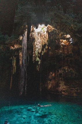 A natural underground pool, known as a cenote, is surrounded by rock formations and illuminated by sunlight streaming through an opening above. Long roots or stalactites hang from the ceiling, creating an otherworldly atmosphere. The water is clear and vibrant turquoise, with a person swimming leisurely in its depths.