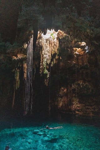 A natural underground pool, known as a cenote, is surrounded by rock formations and illuminated by sunlight streaming through an opening above. Long roots or stalactites hang from the ceiling, creating an otherworldly atmosphere. The water is clear and vibrant turquoise, with a person swimming leisurely in its depths.