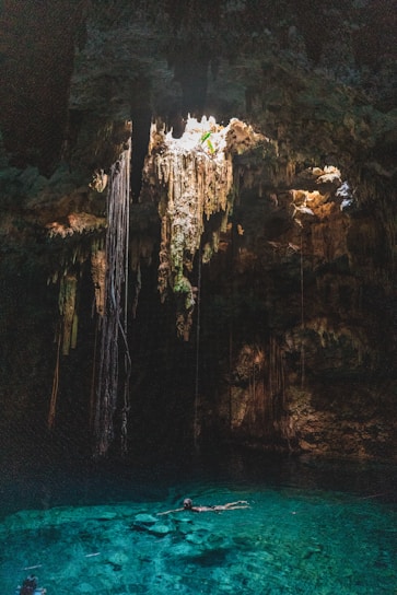A natural underground pool, known as a cenote, is surrounded by rock formations and illuminated by sunlight streaming through an opening above. Long roots or stalactites hang from the ceiling, creating an otherworldly atmosphere. The water is clear and vibrant turquoise, with a person swimming leisurely in its depths.