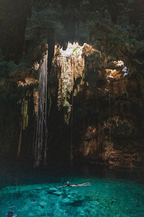 A natural underground pool, known as a cenote, is surrounded by rock formations and illuminated by sunlight streaming through an opening above. Long roots or stalactites hang from the ceiling, creating an otherworldly atmosphere. The water is clear and vibrant turquoise, with a person swimming leisurely in its depths.