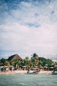 A lively beach scene with several people gathered near the shore, next to thatched-roof huts and lush palm trees. Boats are anchored near the water, and the sky is partially cloudy with sunlight filtering through.