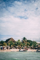 A lively beach scene with several people gathered near the shore, next to thatched-roof huts and lush palm trees. Boats are anchored near the water, and the sky is partially cloudy with sunlight filtering through.