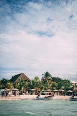 A lively beach scene with several people gathered near the shore, next to thatched-roof huts and lush palm trees. Boats are anchored near the water, and the sky is partially cloudy with sunlight filtering through.