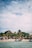 A lively beach scene with several people gathered near the shore, next to thatched-roof huts and lush palm trees. Boats are anchored near the water, and the sky is partially cloudy with sunlight filtering through.