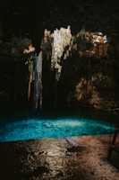 A dimly lit cave with a pool of clear turquoise water at its base. Stalactites hang from the ceiling, illuminated softly by natural light filtering through openings above. The cave walls are rugged and have a mix of dark and earthy tones.