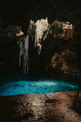 A dimly lit cave with a pool of clear turquoise water at its base. Stalactites hang from the ceiling, illuminated softly by natural light filtering through openings above. The cave walls are rugged and have a mix of dark and earthy tones.