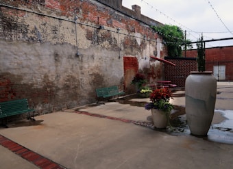 An outdoor courtyard with a worn brick wall creating an aged, rustic atmosphere. Two long green benches sit along the wall, and there's a round red picnic table surrounded by potted plants. A large ceramic pot stands prominently in the foreground, with string lights draping overhead.