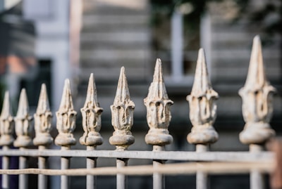 A row of ornate, pointed metal fence finials with a weathered texture, set against a blurred background of a building exterior, creating a sense of depth and focus on the fence.