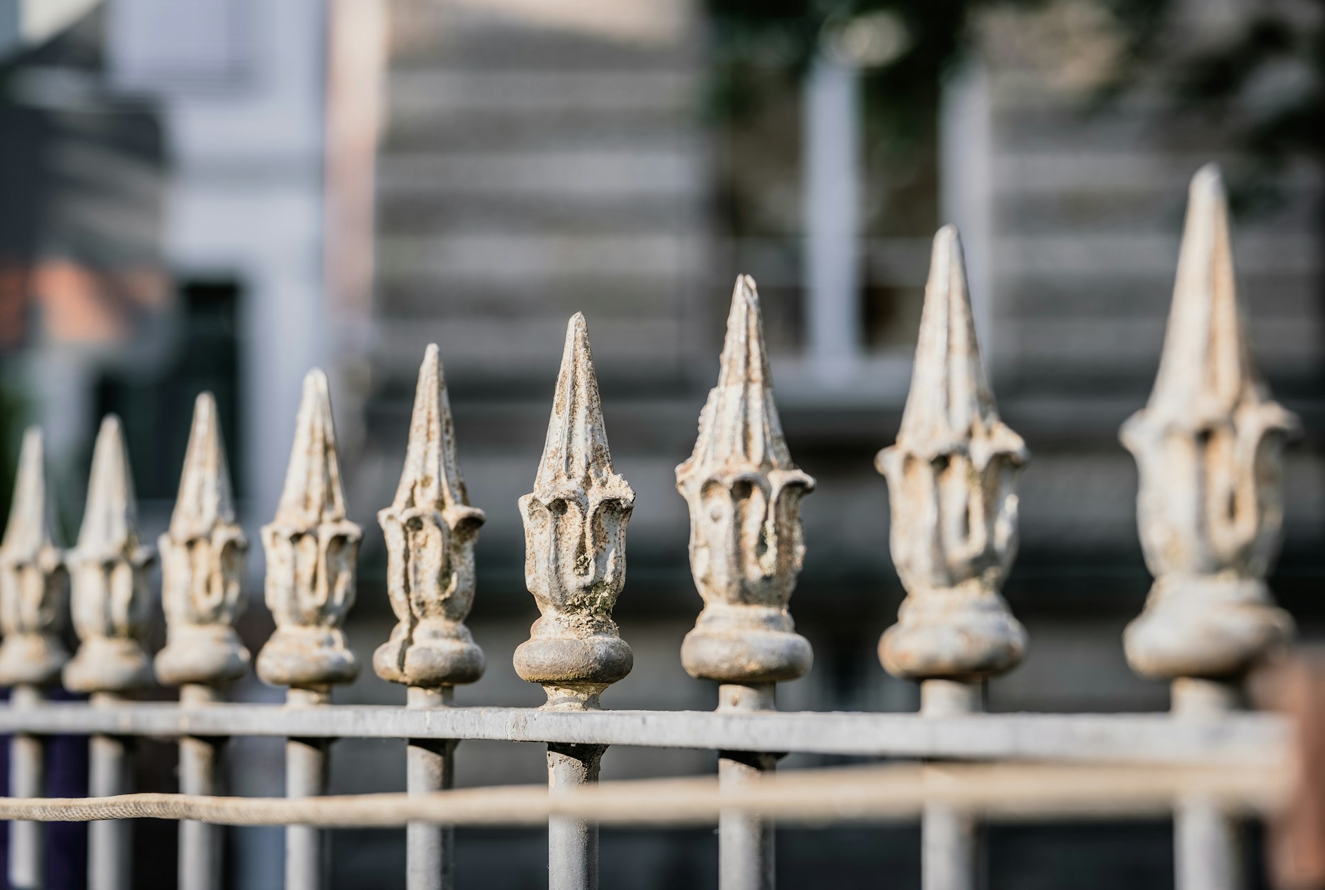 A row of ornate, pointed metal fence finials with a weathered texture, set against a blurred background of a building exterior, creating a sense of depth and focus on the fence.