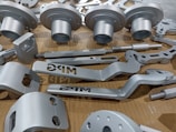 A collection of metallic, machined components neatly arranged on a cardboard surface. The parts vary in shape and size, including cylindrical and flat pieces with holes and grooves. Some parts have the letters 'DRG' etched into them.