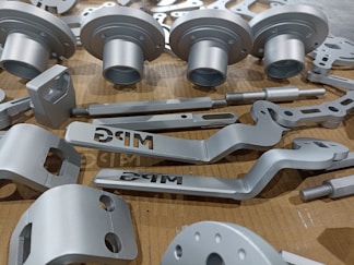 A collection of metallic, machined components neatly arranged on a cardboard surface. The parts vary in shape and size, including cylindrical and flat pieces with holes and grooves. Some parts have the letters 'DRG' etched into them.