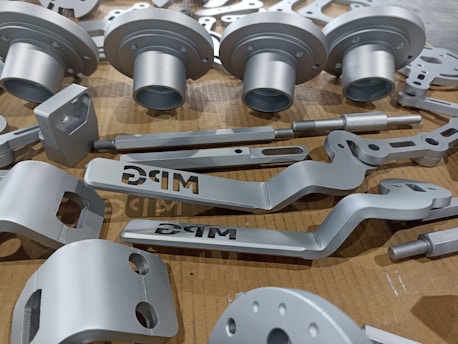 A collection of metallic, machined components neatly arranged on a cardboard surface. The parts vary in shape and size, including cylindrical and flat pieces with holes and grooves. Some parts have the letters 'DRG' etched into them.