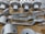 A collection of metallic, machined components neatly arranged on a cardboard surface. The parts vary in shape and size, including cylindrical and flat pieces with holes and grooves. Some parts have the letters 'DRG' etched into them.