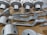 A collection of metallic, machined components neatly arranged on a cardboard surface. The parts vary in shape and size, including cylindrical and flat pieces with holes and grooves. Some parts have the letters 'DRG' etched into them.