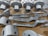 A collection of metallic, machined components neatly arranged on a cardboard surface. The parts vary in shape and size, including cylindrical and flat pieces with holes and grooves. Some parts have the letters 'DRG' etched into them.