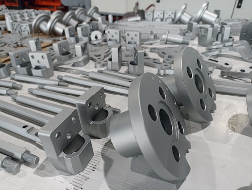 A collection of metallic mechanical parts arranged on a surface, including flanges, shafts, and various fittings with smooth, shiny finishes.