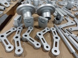 A collection of metallic machine parts arranged on a cardboard surface. The parts include connectors, rods, and brackets, all of which appear to be made of silver-colored metal. The components are neatly laid out, showcasing various shapes and sizes, with some parts having holes for assembly purposes.