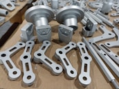A collection of metallic machine parts arranged on a cardboard surface. The parts include connectors, rods, and brackets, all of which appear to be made of silver-colored metal. The components are neatly laid out, showcasing various shapes and sizes, with some parts having holes for assembly purposes.