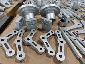 A collection of metallic machine parts arranged on a cardboard surface. The parts include connectors, rods, and brackets, all of which appear to be made of silver-colored metal. The components are neatly laid out, showcasing various shapes and sizes, with some parts having holes for assembly purposes.