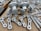 A collection of metallic machine parts arranged on a cardboard surface. The parts include connectors, rods, and brackets, all of which appear to be made of silver-colored metal. The components are neatly laid out, showcasing various shapes and sizes, with some parts having holes for assembly purposes.