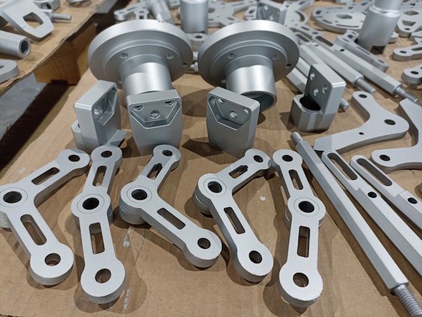 A collection of metallic machine parts arranged on a cardboard surface. The parts include connectors, rods, and brackets, all of which appear to be made of silver-colored metal. The components are neatly laid out, showcasing various shapes and sizes, with some parts having holes for assembly purposes.