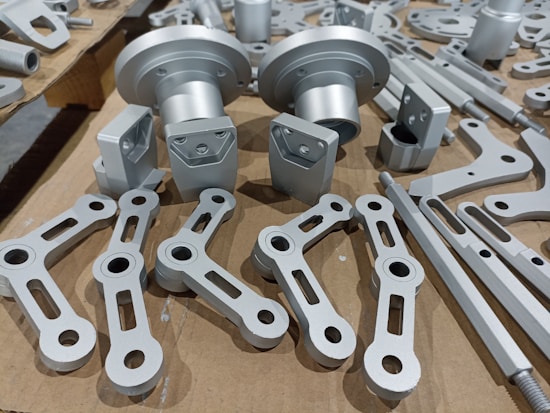 A collection of metallic machine parts arranged on a cardboard surface. The parts include connectors, rods, and brackets, all of which appear to be made of silver-colored metal. The components are neatly laid out, showcasing various shapes and sizes, with some parts having holes for assembly purposes.
