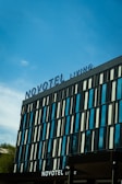 A modern hotel building with a sleek facade featuring vertical glass panels and a large sign displaying 'Novotel' at the top. The sky is clear and blue, with a few clouds visible in the background. There is some greenery visible at the bottom left.
