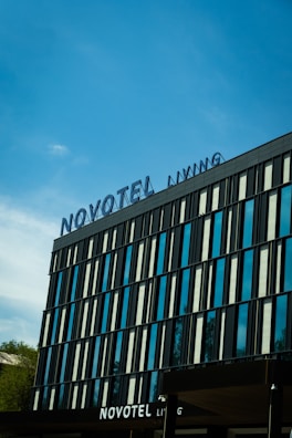 A modern hotel building with a sleek facade featuring vertical glass panels and a large sign displaying 'Novotel' at the top. The sky is clear and blue, with a few clouds visible in the background. There is some greenery visible at the bottom left.