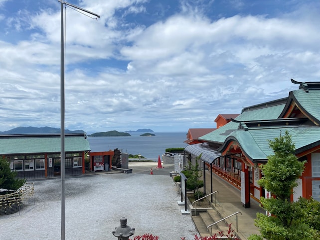 A scenic view featuring traditional Japanese architecture with green rooftops and red accents. The buildings are set against a backdrop of a calm ocean with small islands in the distance. The scene is partially cloudy, with blue skies visible. Greenery, including shrubs and trees, complements the structures.