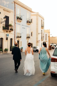 Three people walk down a street beside a light blue building with balconies. The woman in the center is wearing a white dress, possibly a wedding gown, and is accompanied by another woman in a teal dress and a man in a dark suit. The street is lined with cars and plants in pots are visible near the building. The setting appears to be in a town with Mediterranean architecture.