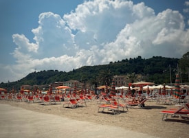 A beach scene featuring numerous orange and green striped umbrellas and sunbeds arranged on sandy ground. In the background, there are lush green hills and a cloudy sky. A few people are lounging under the umbrellas, enjoying the sunny weather.