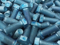 A collection of metal bolts with hexagonal heads and threaded bodies, arranged in a disordered pile. The bolts are predominantly blue with signs of wear and a rough texture.