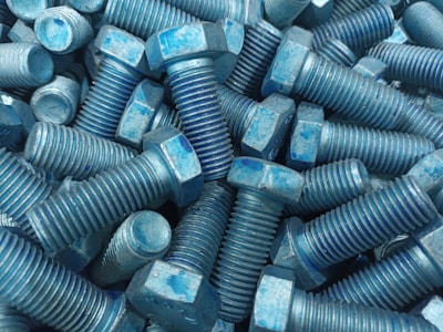 A collection of metal bolts with hexagonal heads and threaded bodies, arranged in a disordered pile. The bolts are predominantly blue with signs of wear and a rough texture.