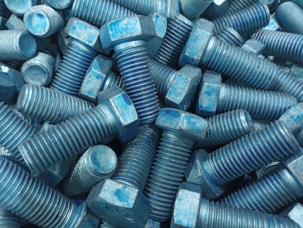 A collection of metal bolts with hexagonal heads and threaded bodies, arranged in a disordered pile. The bolts are predominantly blue with signs of wear and a rough texture.