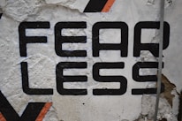 Large bold black typography spelling the word 'FEARLESS' is painted on a rough, textured wall. Some of the paint appears chipped and worn, revealing the underlying surface. There's a hint of orange in a geometric pattern nearby, adding a touch of color.