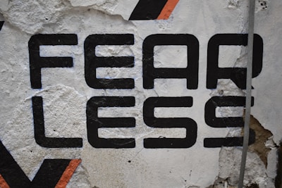 Large bold black typography spelling the word 'FEARLESS' is painted on a rough, textured wall. Some of the paint appears chipped and worn, revealing the underlying surface. There's a hint of orange in a geometric pattern nearby, adding a touch of color.