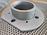 A metallic circular component with multiple holes is placed on a perforated surface. The component has a cylindrical section protruding from a flat base.