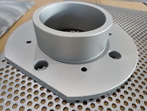 A metallic circular component with multiple holes is placed on a perforated surface. The component has a cylindrical section protruding from a flat base.